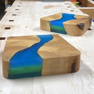 May include: Two wooden coasters with a blue and green epoxy river design. The coasters are made of natural wood and have a smooth, polished finish.