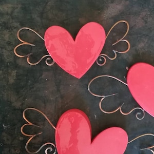 Może przedstawiać: Trzy czerwone drewniane ozdoby w kształcie serca z drucianymi skrzydłami. Serca są pomalowane na jasny czerwony kolor i mają błyszczące wykończenie. Skrzydła są wykonane z cienkiego drutu i są zakręcone na końcach.