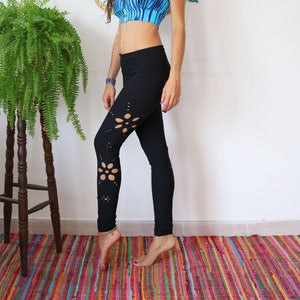 May include: A woman wearing a blue and white striped sports bra and black leggings with floral cutouts. She is standing on a colorful rug in front of a white wall with a mandala design on it.