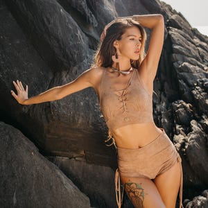 Pode incluir: Uma mulher vestindo um top curto de camurça marrom claro com um cordão na frente e shorts combinando. Os shorts têm um cordão de ajuste nas laterais. Ela está em pé em frente a um fundo rochoso.