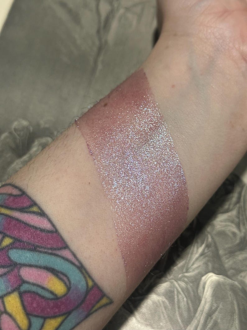 May include: A swatch of shimmering, iridescent makeup applied to the skin. The makeup has a pinkish-mauve base with a sparkling, multi-colored sheen. The swatch is applied in a thick, rectangular shape on the forearm.