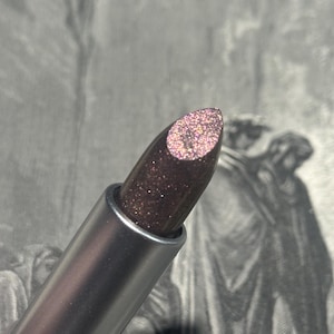 Puede incluir: Primer plano de una barra de labios en un tubo plateado metálico. La barra de labios es de color marrón oscuro con purpurina rosa. El fondo es una ilustración en blanco y negro.
