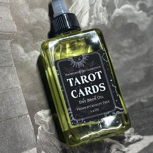 May include: A clear glass bottle with a black label that reads "HAUNTING HUES COSMETICS TAROT CARDS DRY BODY OIL VEGAN & CRUELTY FREE 3.4 Oz". The bottle is filled with a yellow liquid and has a black cap with a clear dropper.