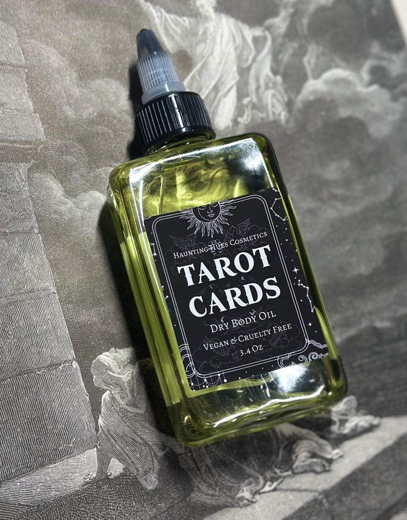 May include: A clear glass bottle with a black label that reads "HAUNTING HUES COSMETICS TAROT CARDS DRY BODY OIL VEGAN & CRUELTY FREE 3.4 Oz". The bottle is filled with a yellow liquid and has a black cap with a clear dropper.