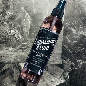 Puede incluir: Una botella de vidrio transparente con una boquilla de pulverización negra y una etiqueta en blanco y negro que dice "Embalm Fluid Makeup Setting Spray Made with Hibiscus Water, Caffeine, Mushroom & Shea Extract 8 oz Vegan & Cruelty Free".