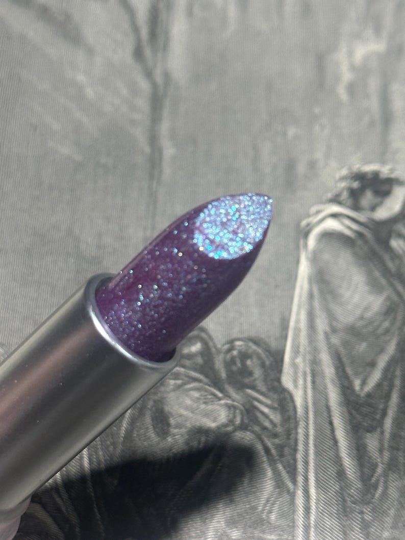 May include: A close-up of a lipstick with a silver tube. The lipstick has a deep purple color with iridescent glitter. The tip of the lipstick has a lighter blue glitter.