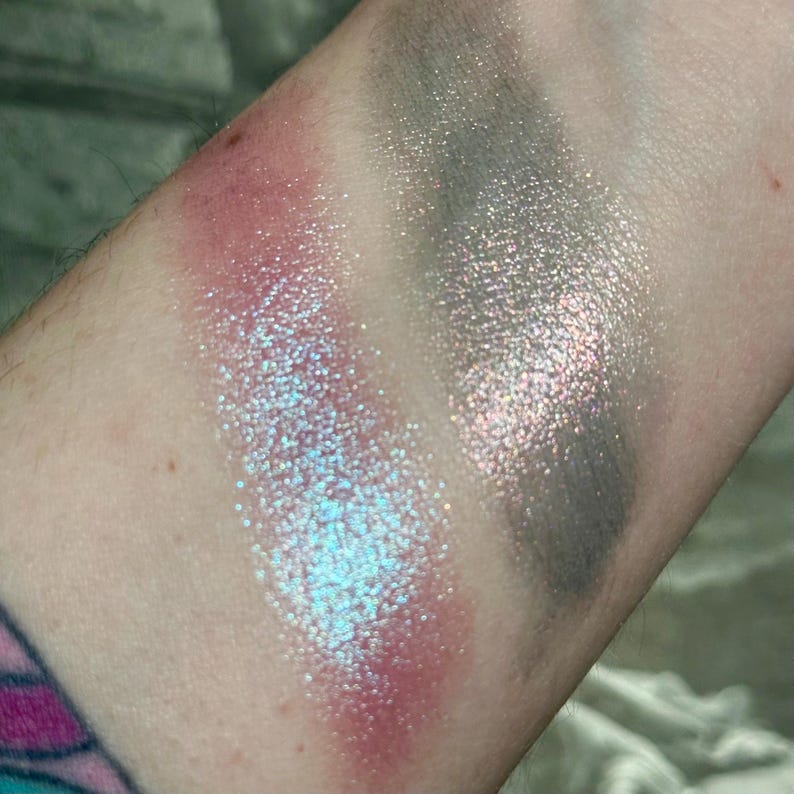 May include: Close-up of three eyeshadow swatches on skin. The first swatch is a shimmery pink, the second is a sparkling blue, and the third is a dark gray with silver glitter. The colors are vibrant and highly pigmented.