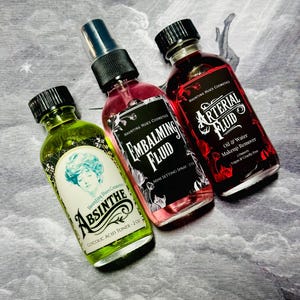 Puede incluir: Tres botellas de vidrio de productos cosméticos con tapas negras. Las botellas están etiquetadas como "Absinthe Glycolic Acid Toner", "Embalming Fluid Mini Setting Spray" y "Arterial Fluid Oil & Water Makeup Remover". Las botellas tienen una estética vintage.