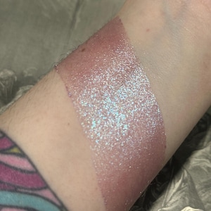 May include: A swatch of shimmering, iridescent pink makeup applied to the skin. The makeup has a glittery, sparkling effect with hints of blue and purple. The color transitions from a darker, more opaque shade to a lighter, more translucent finish.