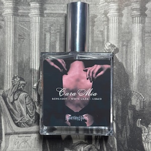 May include: A glass bottle of perfume with a silver cap. The label is black with white text that reads "Cara Mia" and "Bergamot - White Lilies - Amber". The label also includes the text "Haunting Hues".