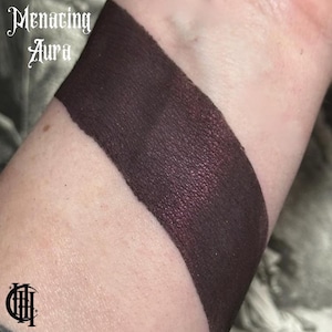 May include: A close-up of a dark purple eyeshadow swatch on a person's arm. The eyeshadow has a shimmery finish and is labelled "Menacing Aura".