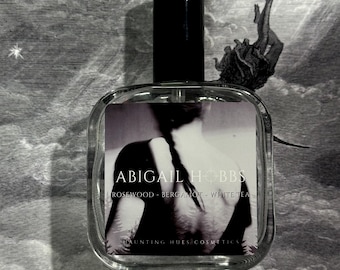 May include: A clear, square perfume bottle with a black cap. The label features the text "ABIGAIL HOBBS" and scent notes. The background is a grayscale illustration with a falling figure and celestial elements. The perfume bottle is the focal point.