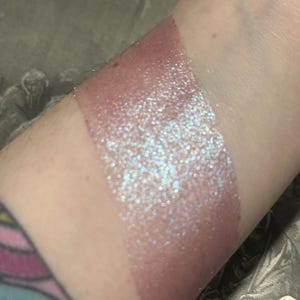 May include: A swatch of shimmering, iridescent makeup on a person's arm. The makeup is a light pink color with a glittery, multi-colored sheen. The swatch is applied in a rectangular shape.