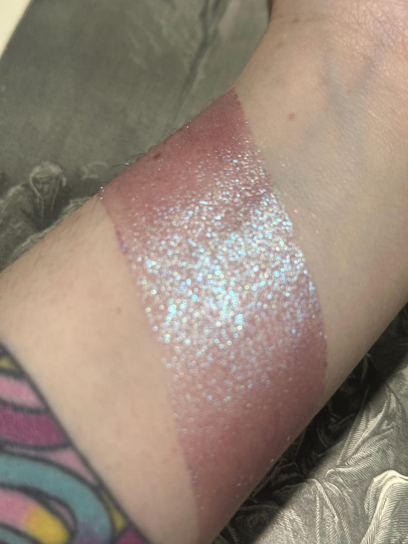 May include: A swatch of shimmering, iridescent makeup on a person's arm. The makeup is a light pink color with a glittery, multi-colored sheen. The swatch is applied in a rectangular shape.