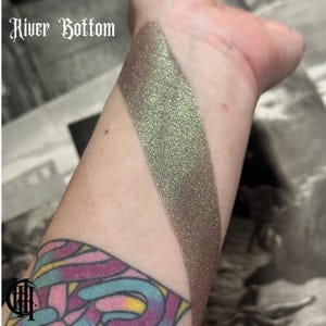 May include: A close-up of a person's arm with a swatch of green and brown eyeshadow with a shimmery finish. The text "River Bottom" is visible at the top of the image.