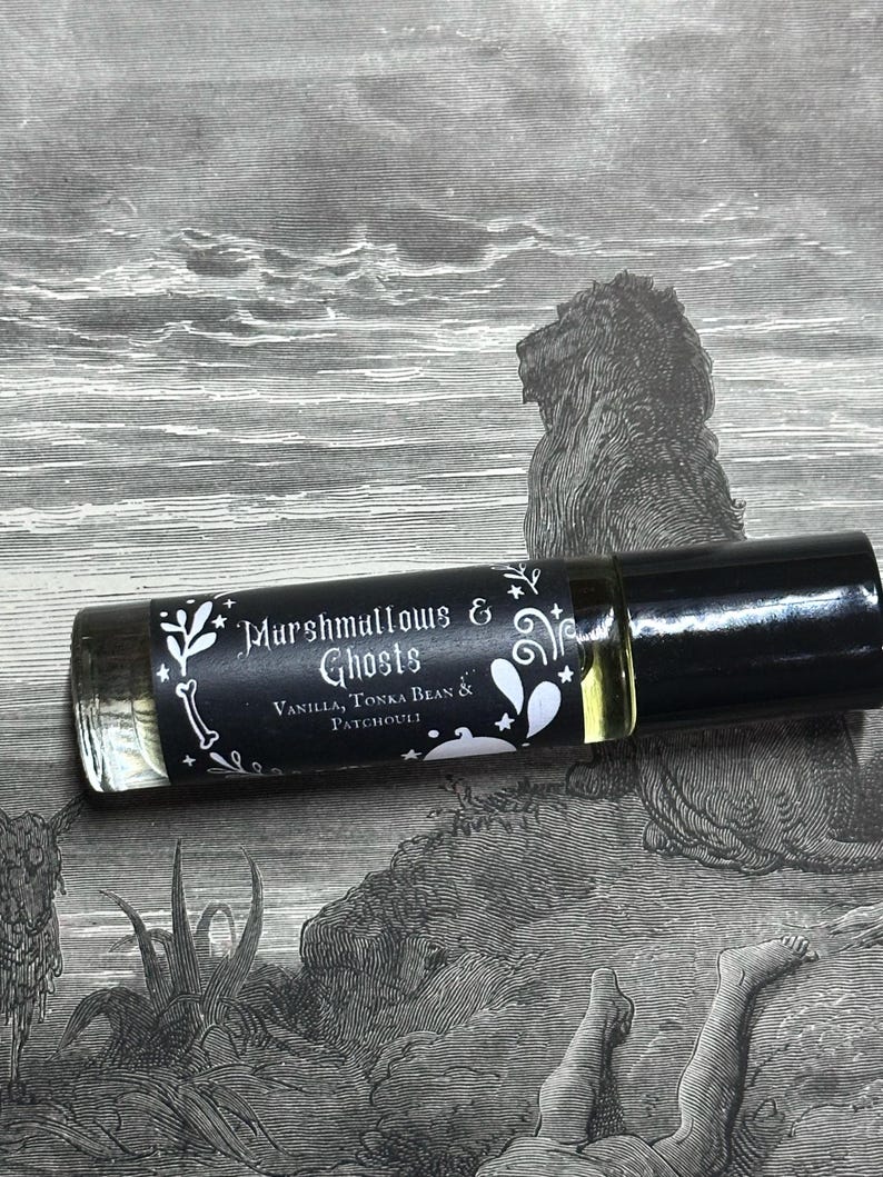 May include: A small, clear glass bottle with a black roll-on top. The label is black with white text that reads "Marshmallows & Ghosts" and "Vanilla, Tonka Bean & Patchouli".