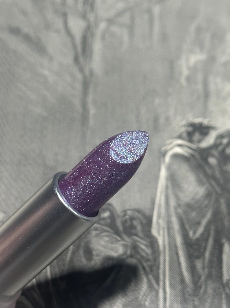 May include: A close-up of a lipstick tube with a silver metallic finish. The lipstick itself is a deep purple color with a glittery, shimmering finish. The tip of the lipstick has a lighter, silver glitter design. The background is a blurred, grayscale image.