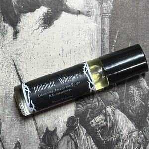 May include: A black and white roll-on bottle of vegan perfume labeled "Midnight Whispers Grave Dirt, Lily of the Valley & Elderflowers Vegan Perfume".