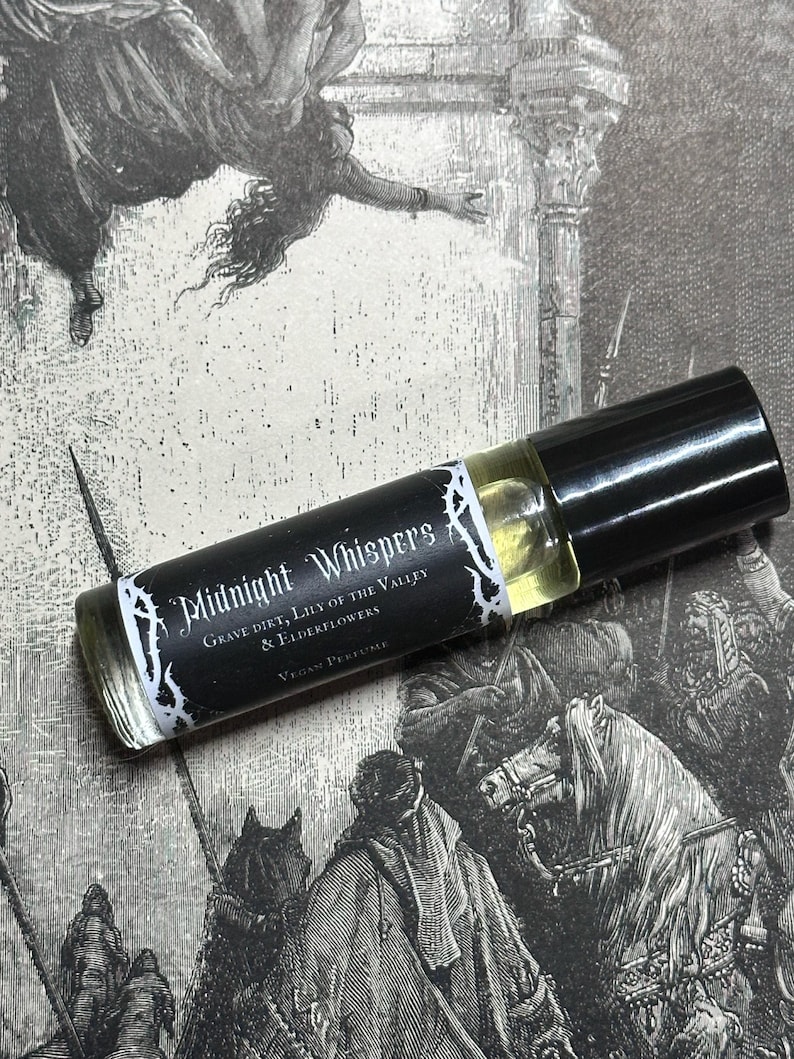 May include: A black and white roll-on bottle of vegan perfume labeled "Midnight Whispers Grave Dirt, Lily of the Valley & Elderflowers Vegan Perfume".