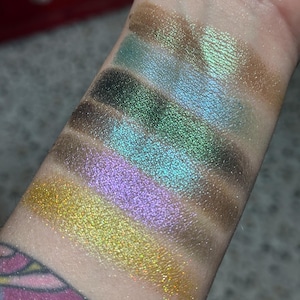 May include: Six horizontal swatches of shimmering eyeshadow applied to skin. The colors include a metallic bronze, a bright teal, a deep forest green, a vibrant turquoise, a soft lavender, and a sparkling yellow gold, all featuring a reflective finish.