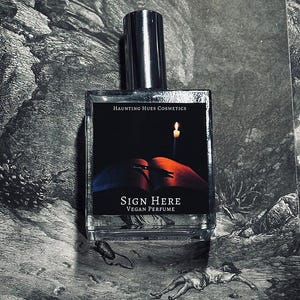 May include: A glass bottle of vegan perfume with a black label that says "Haunting Hues Cosmetics" and "Sign Here Vegan Perfume". The label also features an illustration of a book with a lit candle on top.
