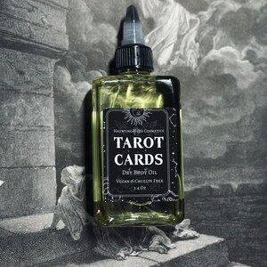 May include: A glass bottle of Tarot Cards Dry Body Oil by Haunting Hues Cosmetics. The bottle is clear and has a black label with white text. The label says "Tarot Cards Dry Body Oil Vegan & Cruelty Free 3.4 oz".