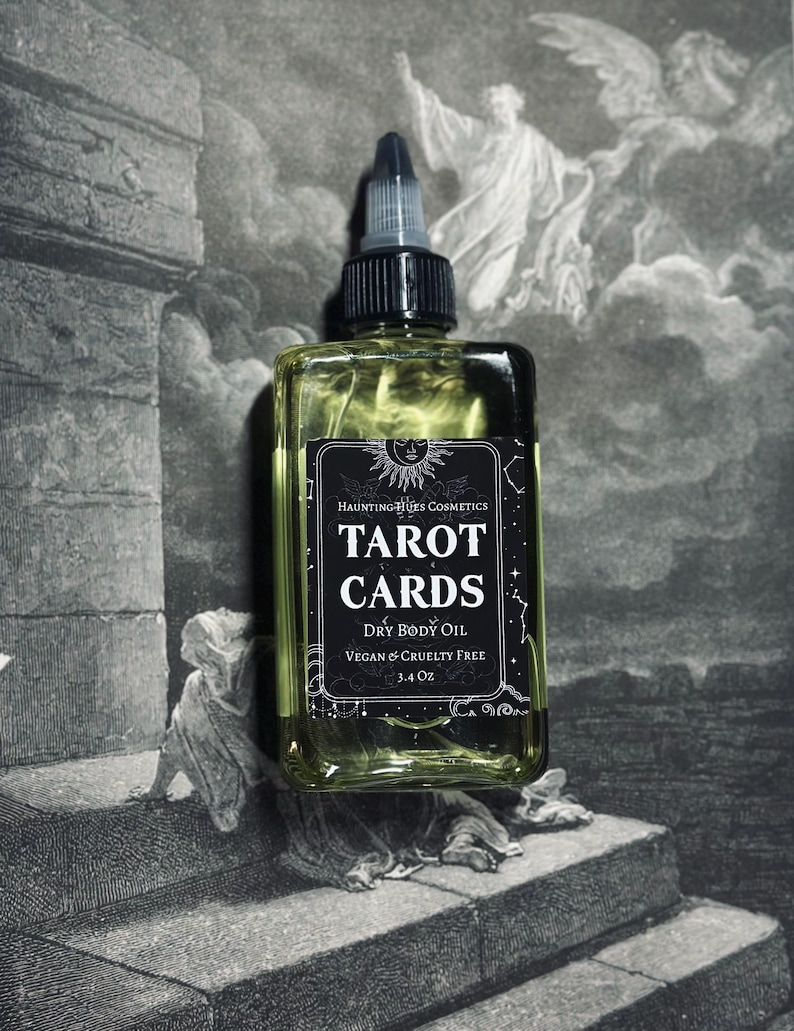 May include: A glass bottle of Tarot Cards Dry Body Oil by Haunting Hues Cosmetics. The bottle is clear and has a black label with white text. The label says "Tarot Cards Dry Body Oil Vegan & Cruelty Free 3.4 oz".