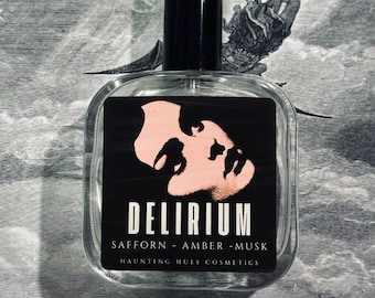May include: A clear glass perfume bottle with a black cap. The label features the word "DELIRIUM" in white, with "SAFFORN - AMBER - MUSK" below. A pink face graphic is above the word "DELIRIUM". The background is a black and white illustration.
