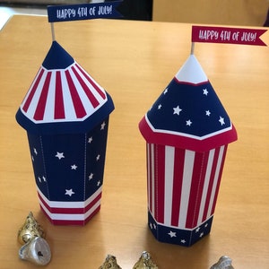 Könnte beinhalten: Zwei rot-weiß-blaue Papier-Party-Geschenkboxen in Form von Feuerwerkskörpern mit dem Text "Happy 4th of July!" auf einer Flagge auf jeder Box. Es gibt sechs goldfolienverpackte Schokoladenküsse auf einer Holzoberfläche.