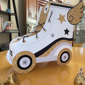 May include: A white and gold roller skate party favour box with a black star and a gold glitter wing. The box has a black and white wheel with a gold glitter rim.