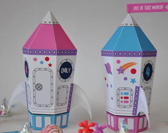May include: Two paper rocket party favor boxes, one pink and one blue, with space-themed designs. The pink rocket has the name "Emily" on it, and the blue rocket has the number "4" on it. The blue rocket has a banner that says "Out of this world!"