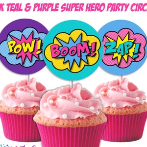 Könnte beinhalten: Drei rosa Cupcakes mit weißem Frosting und Streuseln, die mit runden Cupcake-Toppern im Superhelden-Design verziert sind. Die Topper sind lila, türkis und blau mit gelbem, rosa und türkisfarbenem Text, der "POW!" "BOOM!" und "ZAP!" lautet.