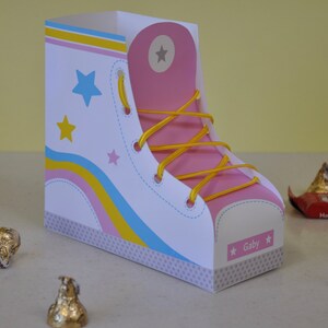 Peut inclure: Une bo&icirc;te en forme de chaussure blanche et rose avec des lacets jaunes et des rayures bleues et jaunes. La bo&icirc;te a un motif d'&eacute;toile et le nom "Gaby" sur le c&ocirc;t&eacute;.