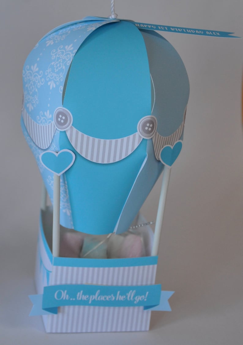 May include: A blue and white paper hot air balloon with a banner that reads "Oh... the places he'll go!" and a smaller banner that reads "Happy 1st Birthday Alex".