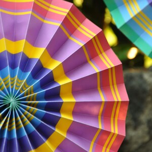 K&ouml;nnte beinhalten: Ein bunter Papierf&auml;cher mit lila, rosa, gelben und blauen Streifen. Der F&auml;cher ist kreisf&ouml;rmig gefaltet und hat einen Mittelpunkt.