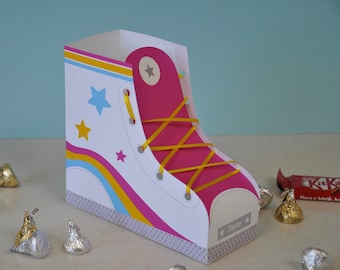 Può includere: Una scatola a forma di scarpa bianca e rosa con lacci di nastro giallo e un design blu, giallo e rosa. La scatola ha un motivo a stelle e il nome "Taylor" scritto sul fondo.
