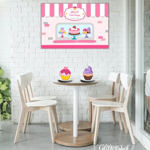 May include: A pink and white bakery storefront backdrop with a sign that reads "Emily's Bake Shoppe". There are two cupcakes on a table in front of the backdrop, one with pink frosting and one with purple frosting.