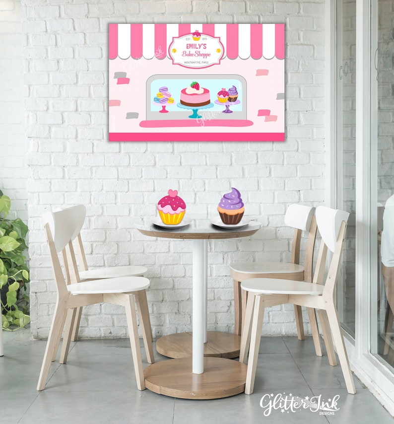 May include: A pink and white bakery storefront backdrop with a sign that reads "Emily's Bake Shoppe". There are two cupcakes on a table in front of the backdrop, one with pink frosting and one with purple frosting.