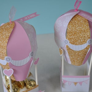 May include: Two pink and gold hot air balloon party favours with white and pink striped baskets. The balloons have gold glitter and white patterned paper. One balloon has a banner that says "Up, Up & Away" and the other says "Happy First Birthday".