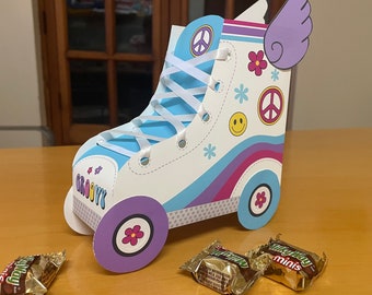 Può includere: Una scatola portaconfetti a forma di pattini a rotelle in cartone blu e viola con un nastro bianco e le parole "Groovy" sul lato. La scatola ha un'ala viola sulla parte superiore e un segno di pace sul davanti.