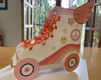 Può includere: Un pattino a rotelle di cartone bianco e rosa con lacci di nastro arancione e un design a forma di segno della pace. Il pattino ha un'ala rosa sul lato e un'emoticon sorridente sul davanti.