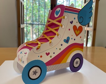 Può includere: Una scatola di cartone per bomboniere a forma di pattino a rotelle, decorata con un arcobaleno, cuori e una stella sulle ruote. La scatola è decorata con un nastro giallo e il nome "Sophia" è scritto sul lato.