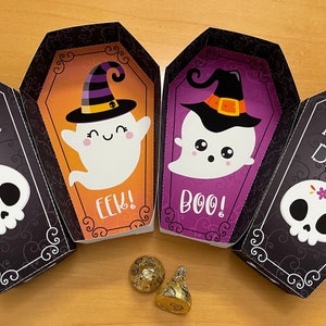 Puede incluir: Cuatro cajas en forma de ataúd negras y blancas con una calavera y el texto "Trick or Treat!" en la parte delantera. Las cajas están abiertas para revelar un fantasma con un sombrero de bruja y el texto "Boo!" y "Eek!" en el interior. Dos monedas de chocolate doradas están delante de las cajas.