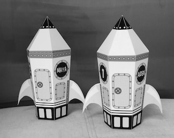 May include: Two white cardboard rocket-shaped party favor boxes with black and gray details. The boxes have cut-out windows with the names "MAYA" and "AIDEN" written on them.