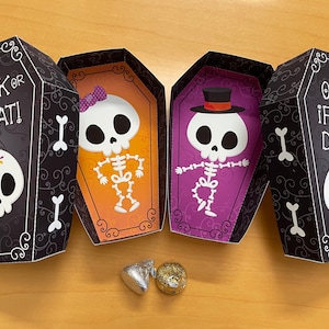 Puede incluir: Dos cajas negras y blancas en forma de ataúd con personajes de esqueleto en su interior. Las cajas tienen el texto "Trick or Treat!" y "Open if you dare!" en ellas. Las cajas están abiertas y hay dos caramelos envueltos en plata y oro en su interior.