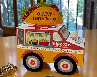 Può includere: Un camioncino per tacos di cartone rosso, giallo e bianco con le parole "Olivia's Fiesta Tacos" in cima. Il camioncino ha una finestra con le parole "tacos nachos * burritos" su di essa. C'è una piccola barretta di cioccolato in primo piano.