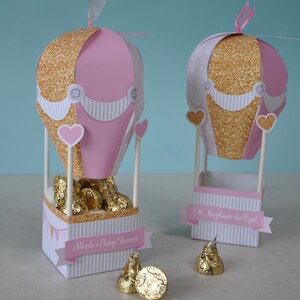 May include: Two pink and gold hot air balloon party favour boxes with white and gold striped bases. The boxes are filled with chocolate kisses and have a banner that says "Oh... the places she'll go!" and "Nicole's Baby Shower".