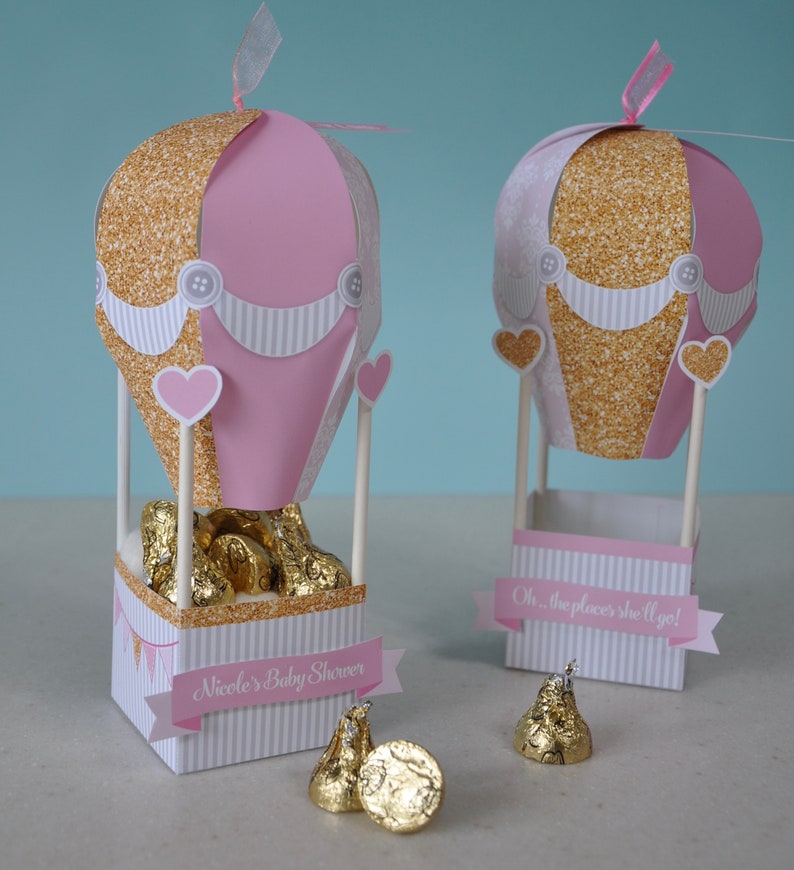 May include: Two pink and gold hot air balloon party favour boxes with white and gold striped bases. The boxes are filled with chocolate kisses and have a banner that says "Oh... the places she'll go!" and "Nicole's Baby Shower".