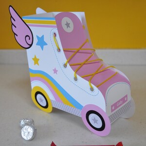 Peut inclure: Une bo&icirc;te &agrave; drag&eacute;es en forme de patin &agrave; roulettes en carton rose et blanc avec des lacets jaunes, des rayures bleues et jaunes et des &eacute;toiles. La bo&icirc;te a une aile rose sur le c&ocirc;t&eacute; et le nom "Gaby" imprim&eacute; sur le devant.