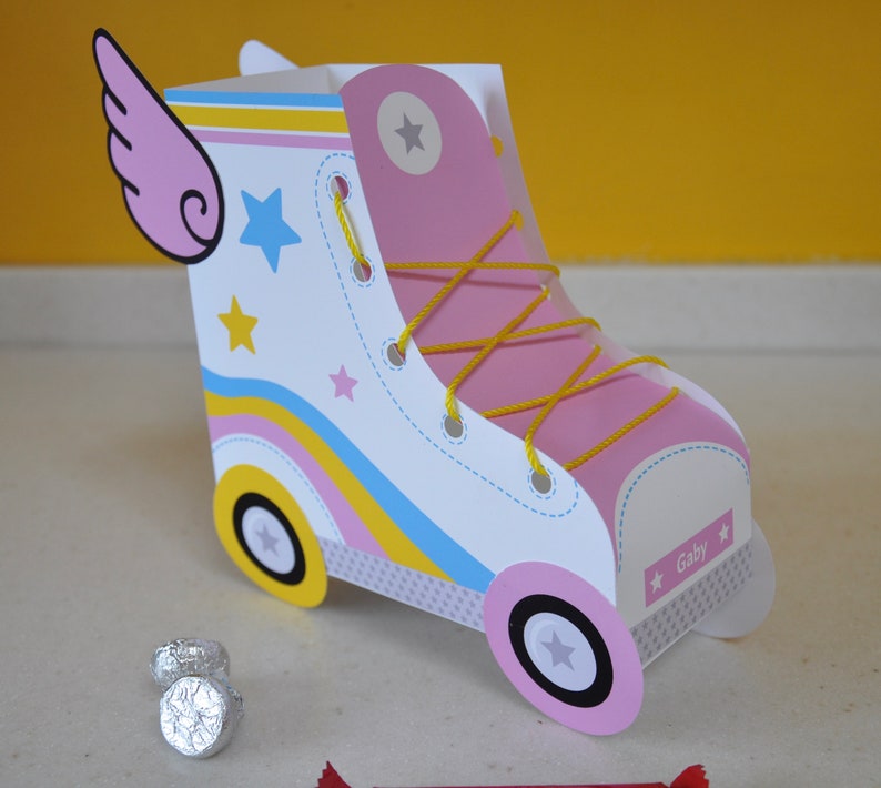 Peut inclure: Une bo&icirc;te &agrave; drag&eacute;es en forme de patin &agrave; roulettes en carton rose et blanc avec des lacets jaunes, des rayures bleues et jaunes et des &eacute;toiles. La bo&icirc;te a une aile rose sur le c&ocirc;t&eacute; et le nom "Gaby" imprim&eacute; sur le devant.